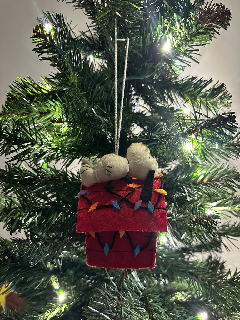 Tree ornament of a white dog laying on top of a red dog house decorated in rainbow Christmas lights.