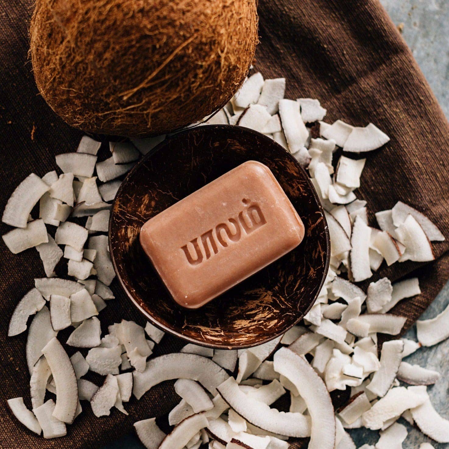 Bar of Unown soap in a coconut shell surrounded by coconut pieces on a wooden surface.