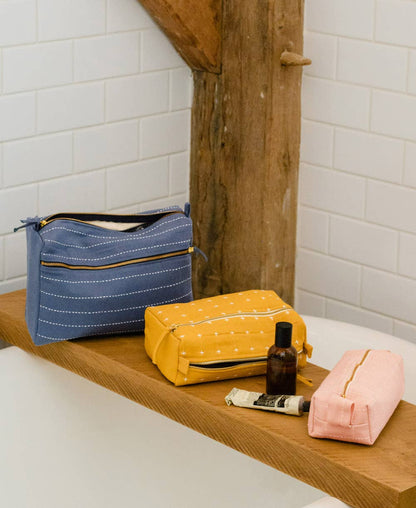 Three cosmetic bags in blue, yellow, and pink on a wooden shelf with bathroom tiles in the background.