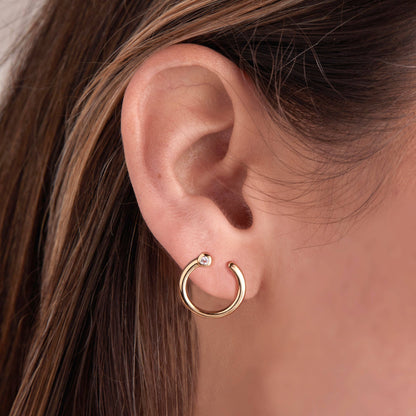 Close-up of an ear wearing a gold hoop earring with a clear gemstone.