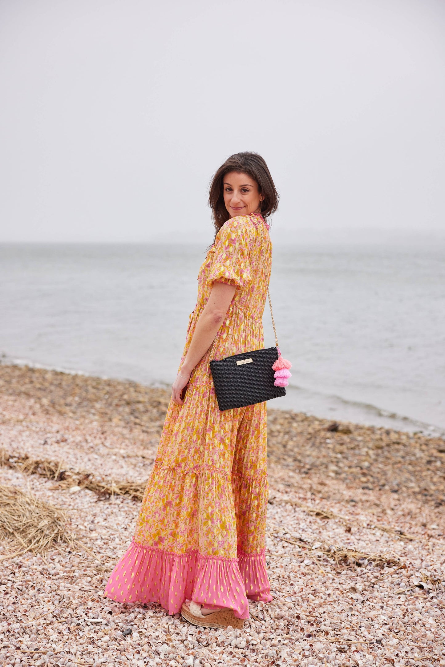 Woman in a yellow floral dress with pink hem standing on a pebbly beach.