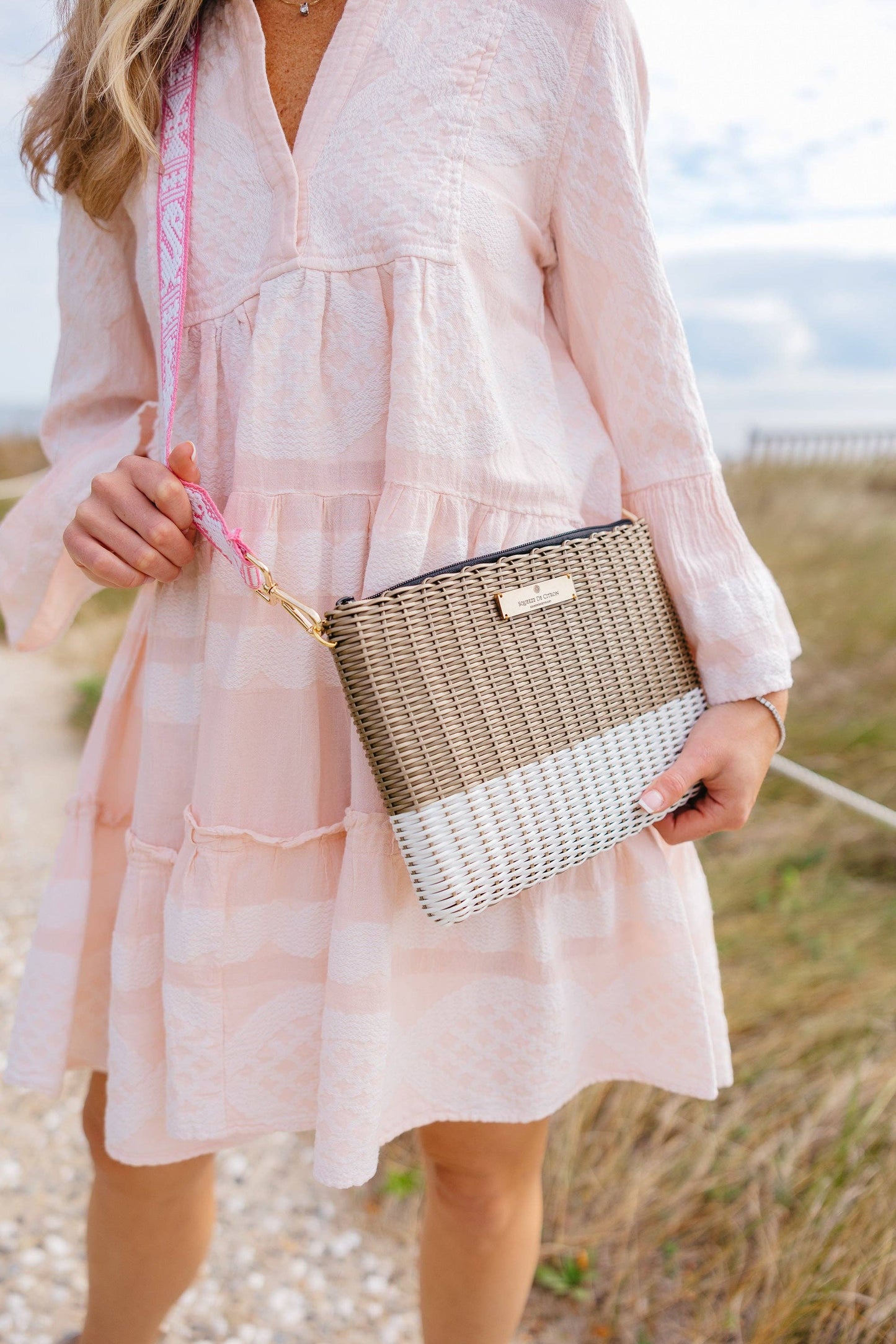 Woman in a light pink dress holding a woven handbag in an outdoor setting.