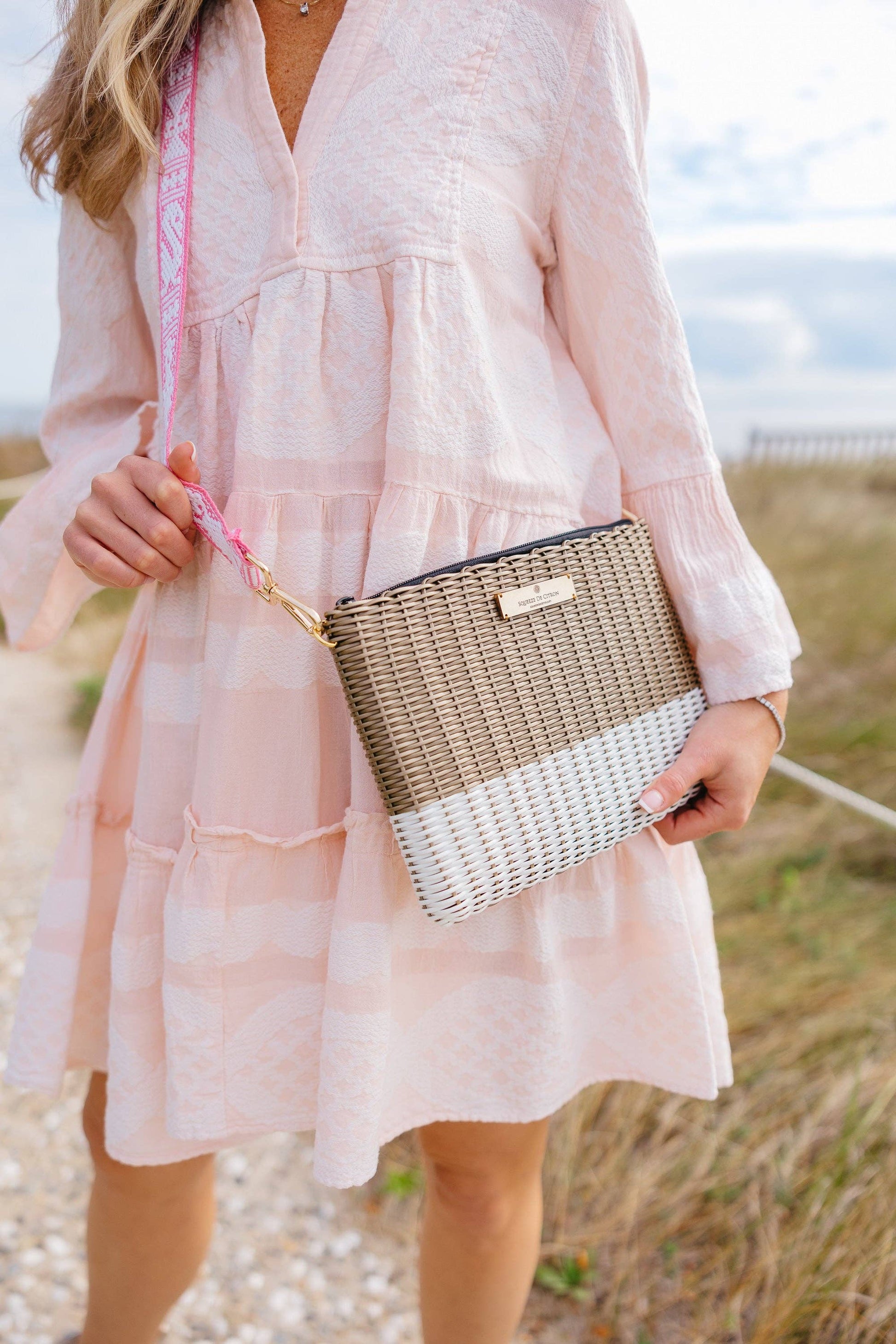 Woman in a light pink dress holding a woven handbag in an outdoor setting.