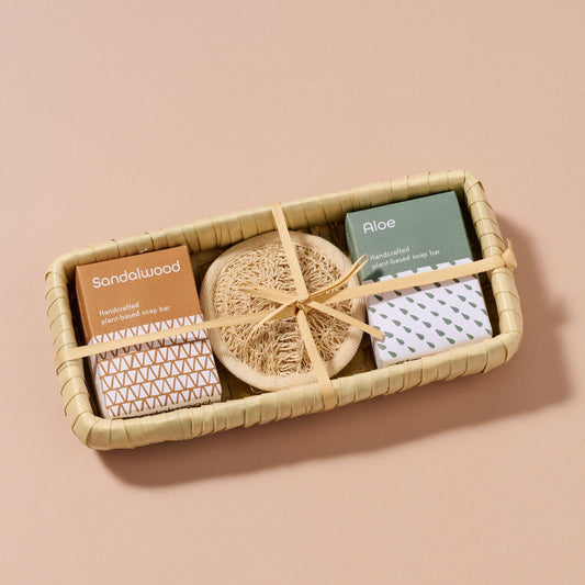Set of bath products including a bar of soap, loofah, and washcloth in a woven tray on a beige background.