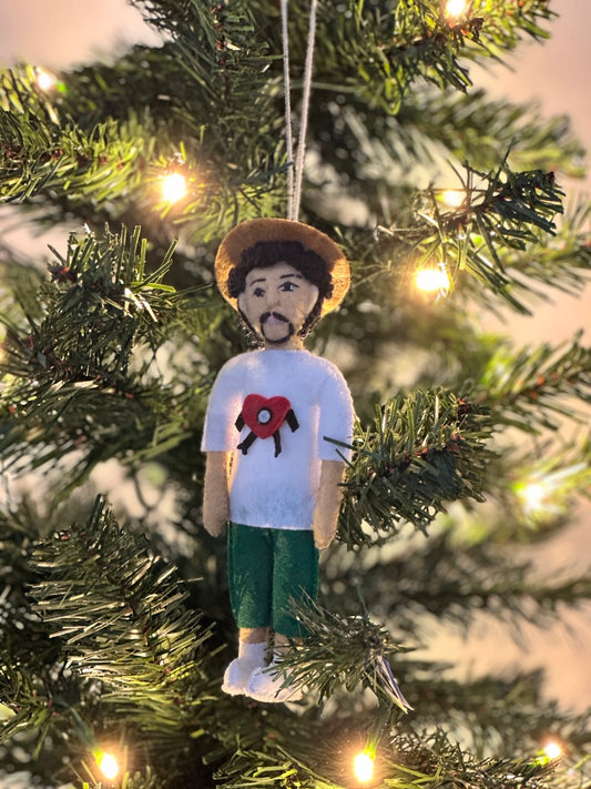 Ornament of a person wearing a white shirt with a red design, green shorts, and a hat, hanging on a Christmas tree.