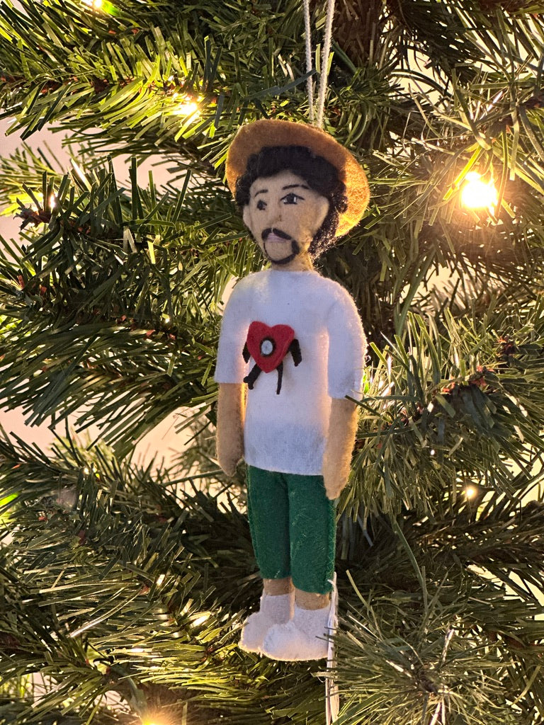 Ornament of a person wearing a white shirt with a red heart design, green shorts, and a brown hat, hanging on a Christmas tree.