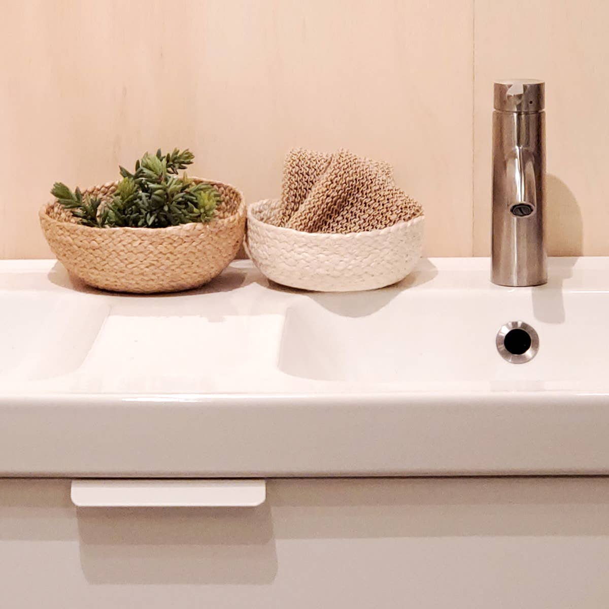 Bathroom sink with two woven baskets, one with plants and the other with a towel, next to a metallic faucet.