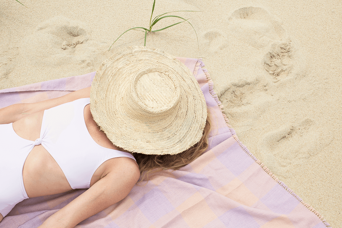 Person lying on a beach towel wearing a white swimsuit and straw hat.