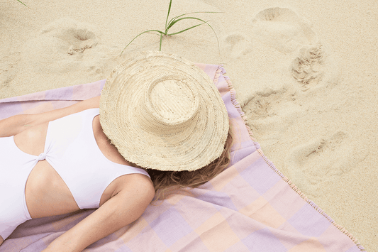 Person lying on a beach towel wearing a white swimsuit and straw hat.