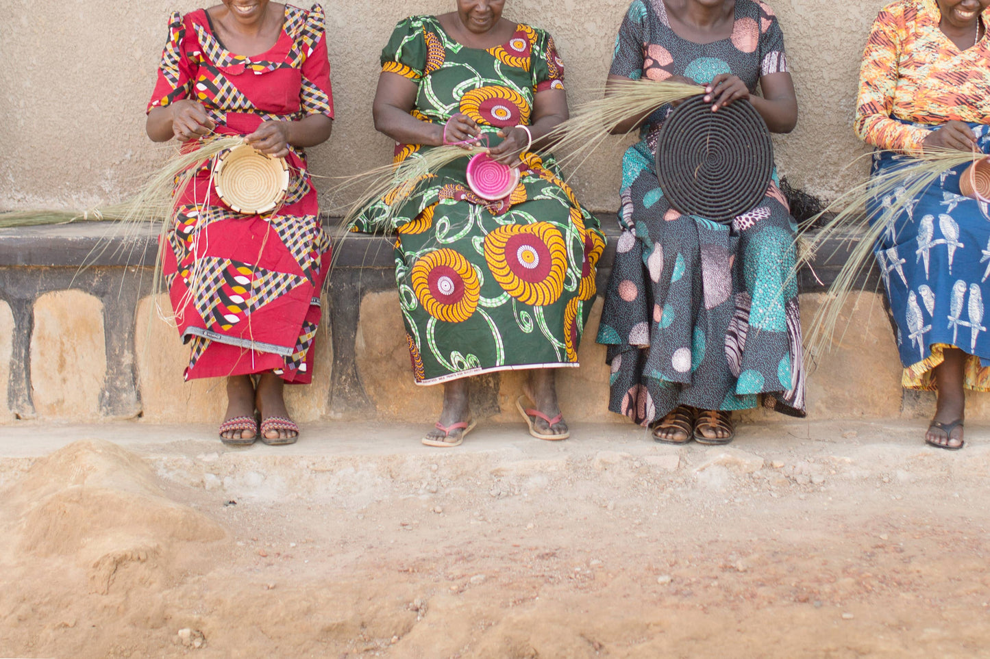 Four women sitting on a stone wall, each holding a woven basket, with a rustic background.