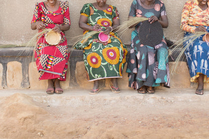 Four women sitting on a stone wall, each holding a woven basket, with a rustic background.