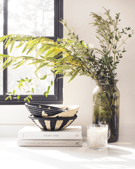 Decorative setup with books, a striped bowl, a vase with greenery, and a candle on a white surface.