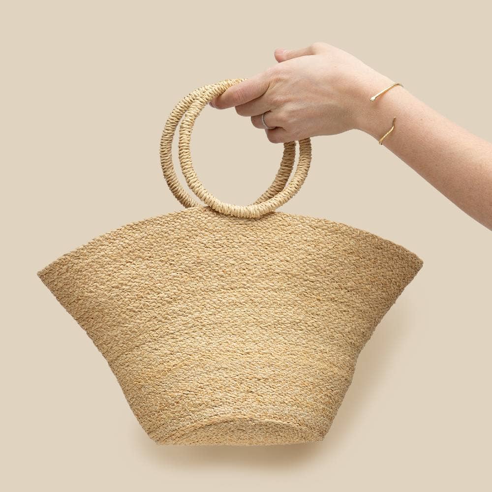 Hand holding a woven beige handbag with circular handles against a beige background