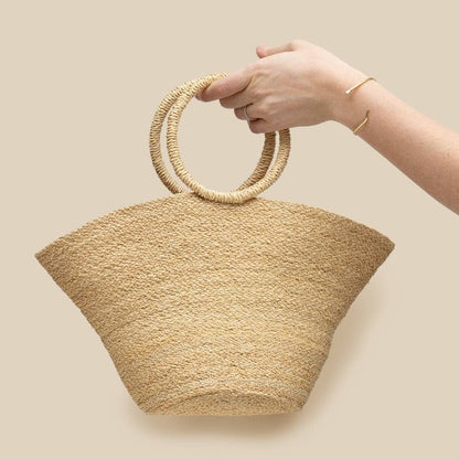 Hand holding a woven beige handbag with circular handles against a beige background