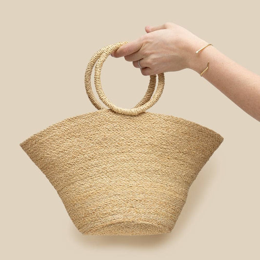 Hand holding a woven beige handbag with circular handles against a beige background