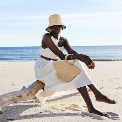 Woman sitting on a beach with a hat and bag, wearing a white dress.