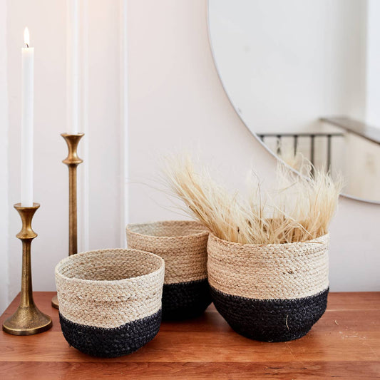 Three woven baskets with black bottoms on a wooden surface, with candles and a mirror in the background.