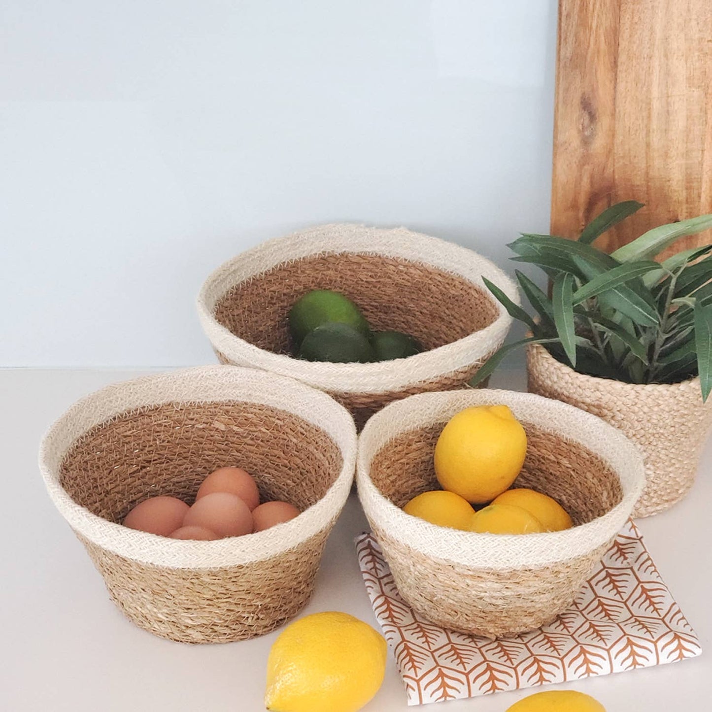 Three woven baskets with eggs, lemons, and a plant on a light surface.