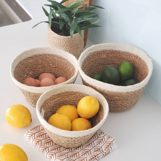 Three woven bowls containing eggs, lemons, and limes on a white surface with a plant in the background.