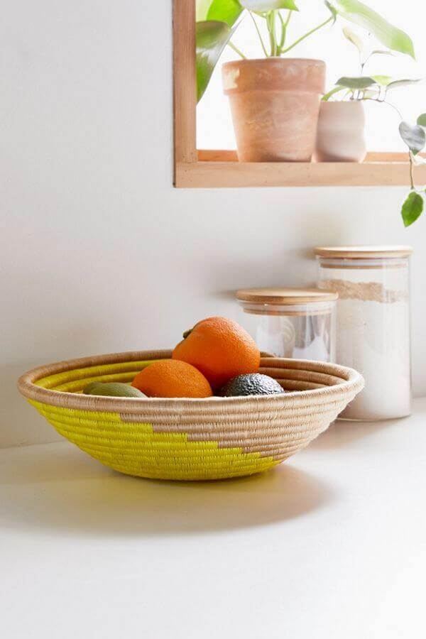 Woven fruit bowl with oranges and avocados on a white surface, with jars and a plant in the background.